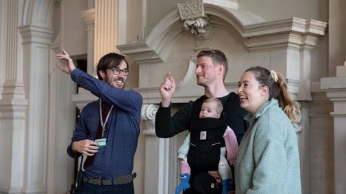 A room guide talking to male and female visitors, the man has a baby in a baby carrier on his front, in the Great Hall at Beningbrough, North Yorkshire